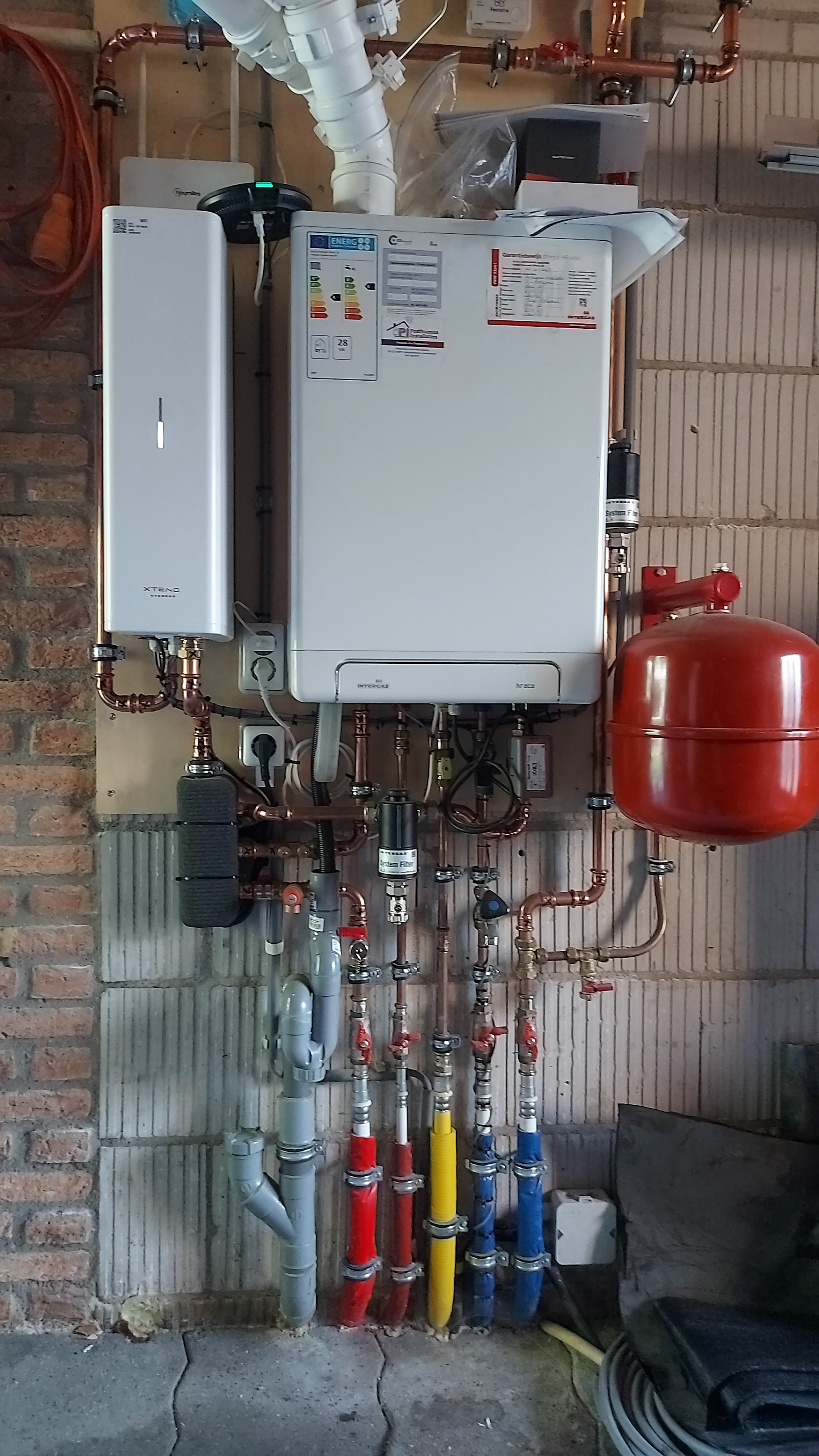 Nieuwe cv-ketel en boiler aan een bakstenen muur, met een rood expansievat en rood, blauw en geel gemerkte leidingen